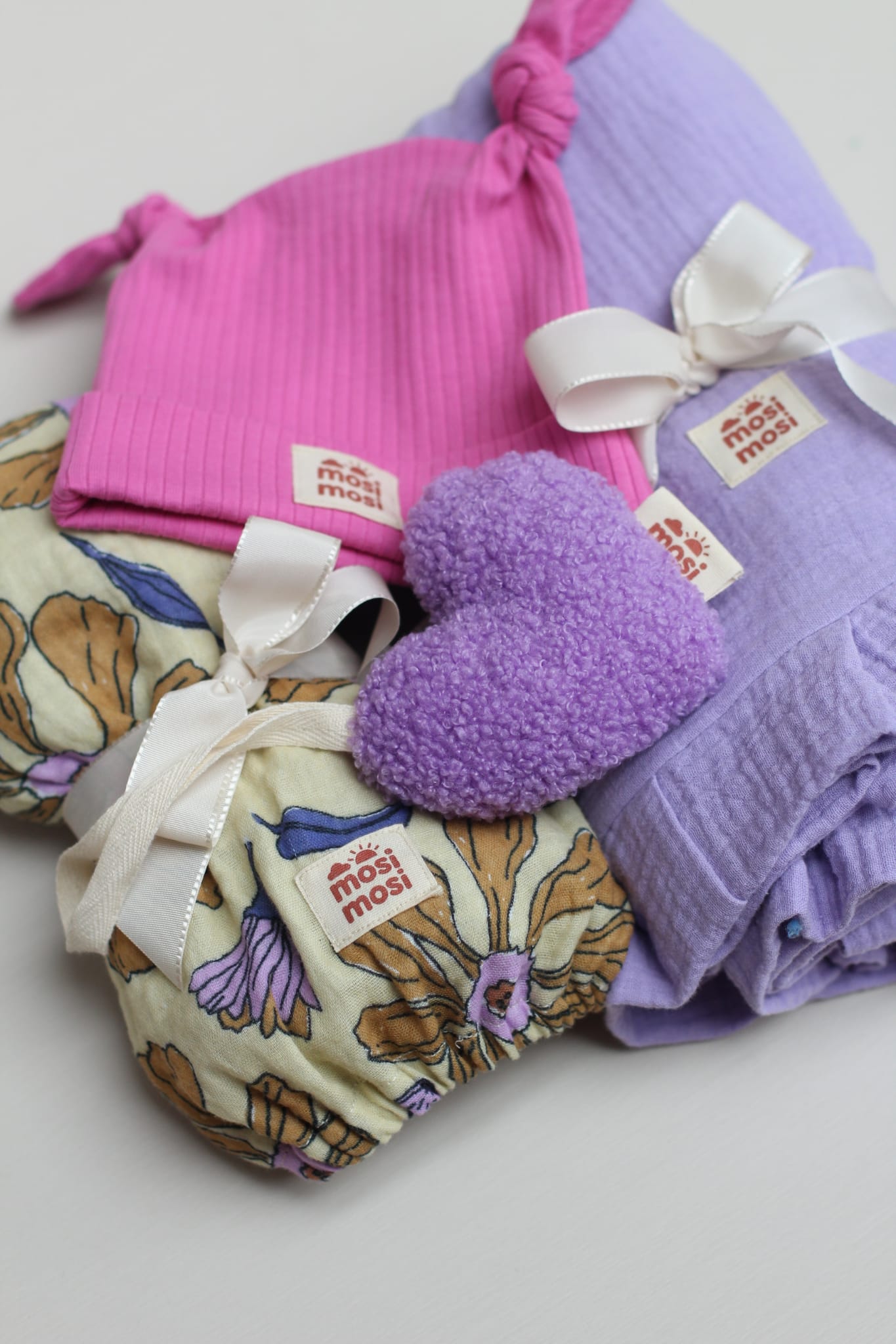 A set of products to be used as gift, showing a pink hat, purple blanket, purple heart-shaped plush-toy and colourful fitted sheet
